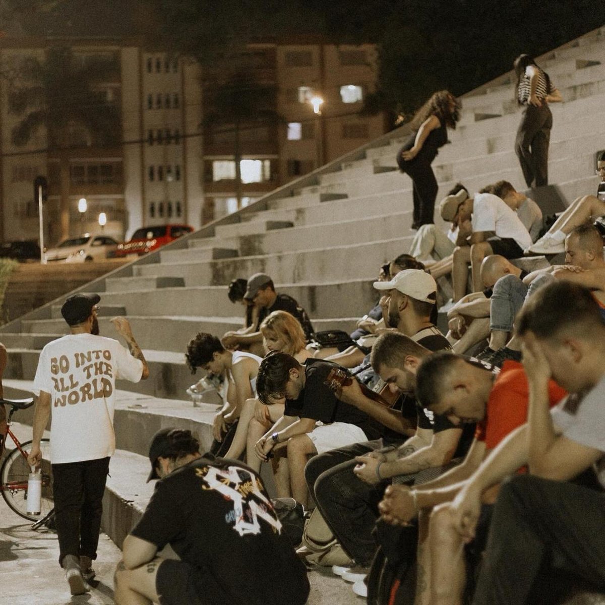 Matheus caminhando entre jovens que oram sentados na escadaria de uma praça à noite